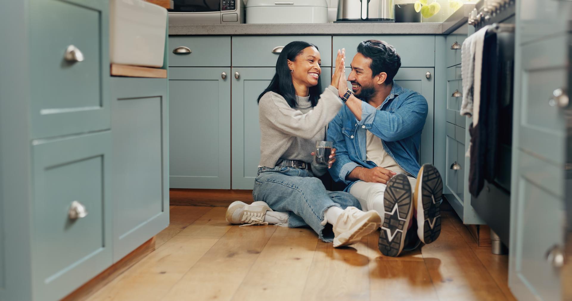 Pareja, café y sentarse en el suelo de la cocina para pasar tiempo de calidad o divertirse para celebrar o discutir. El amor, la sonrisa y la mujer choca los cinco se unen con la cafeína y la conversación o el cuidado.