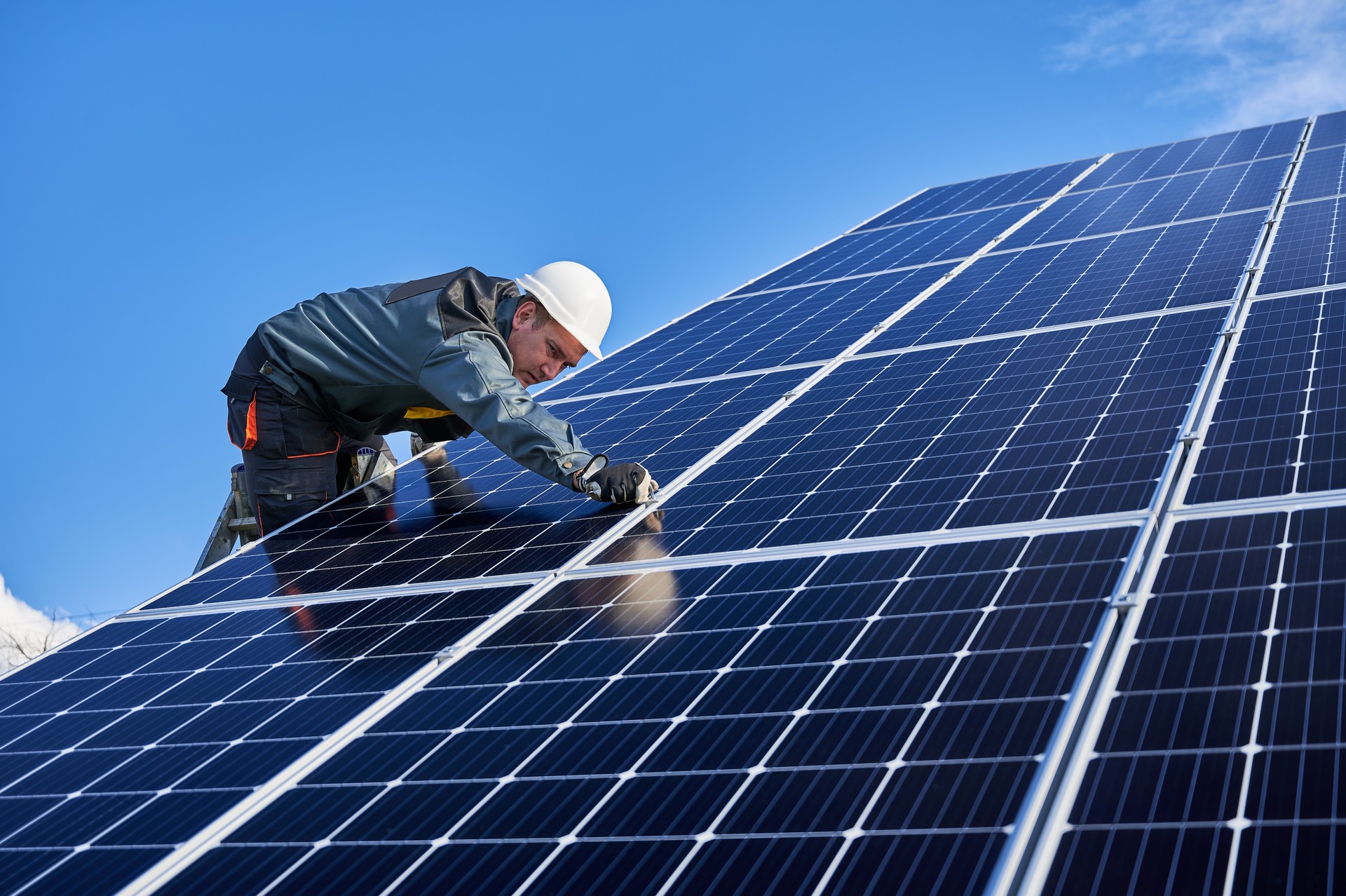 Trabajador masculino en la planta solar instalando baterías solares, de pie en una escalera en un día soleado