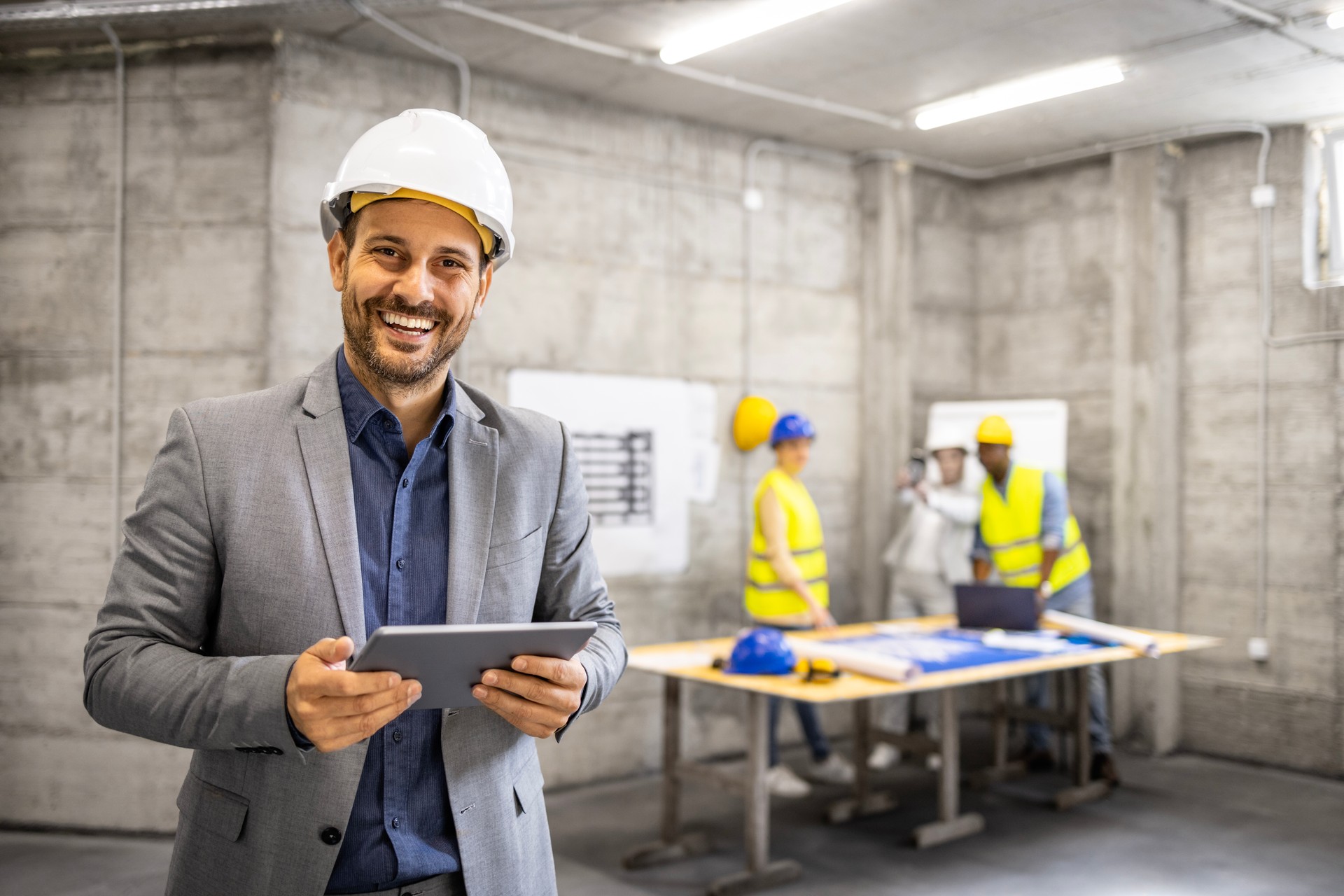 Retrato de un arquitecto experimentado con casco sosteniendo una tableta digital mientras los ingenieros civiles planean en segundo plano.